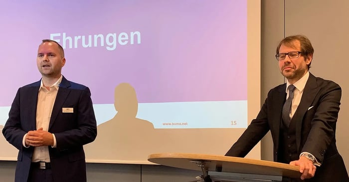 Lars Müller and Michael Kulig stand in front of a screen showing the word 'Ehrungen' (Honors) during the BVMS General Assembly, leading the official awards segment.