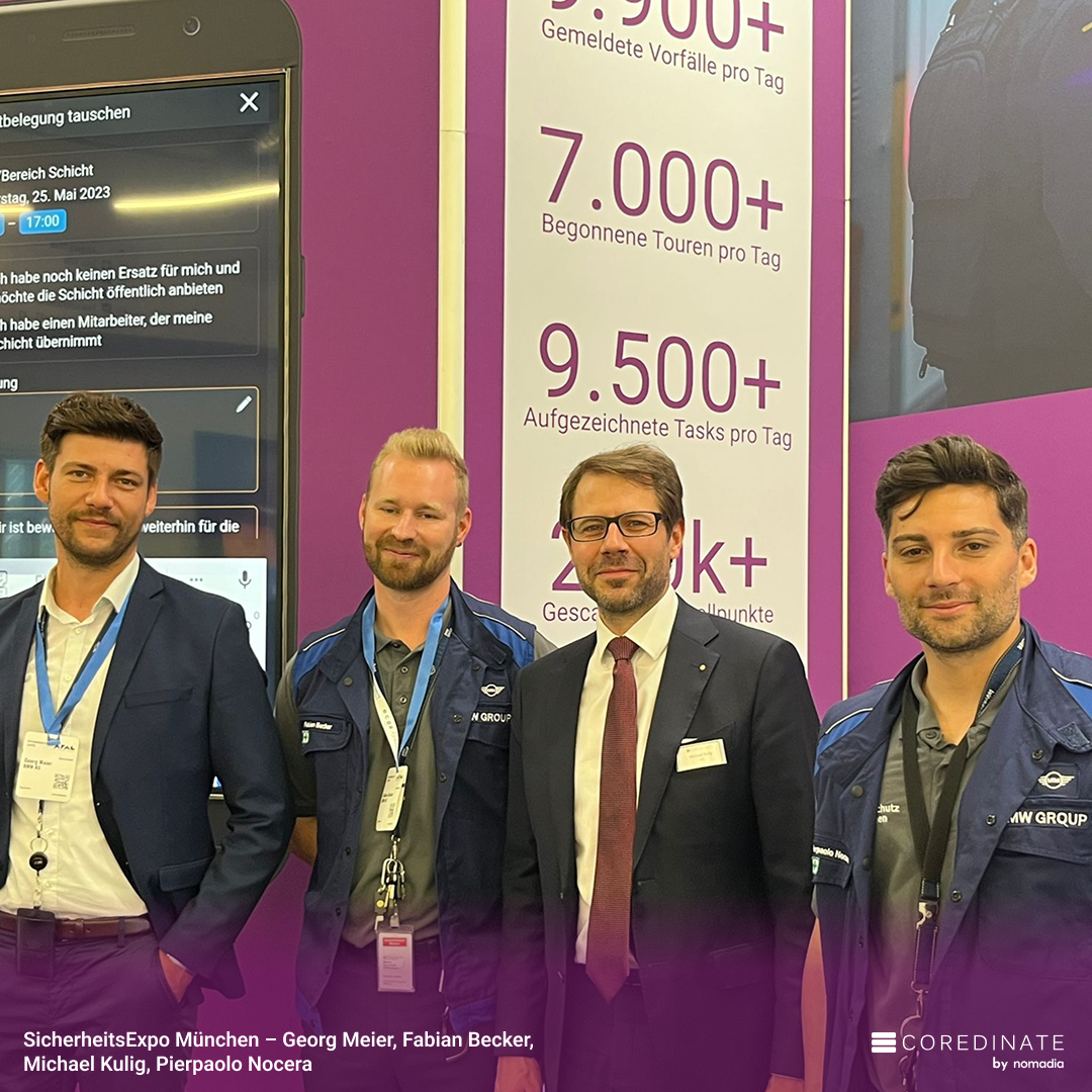 Quatre hommes (Georg Meier, Fabian Becker, Michael Kulig, Pierpaolo Nocera) souriant pour une photo de groupe à la SecurityExpo Munich, devant le stand violet COREDINATE® avec des statistiques d’activités.