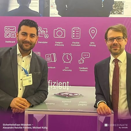 Alexandre Reiche Ferreira and Michael Kulig standing next to each other at the COREDINATE® booth at SecurityExpo Munich, smiling at the camera.
