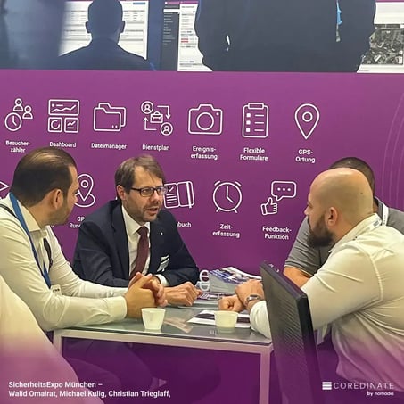 Four men having a discussion at a table at SecurityExpo Munich, including Walid Omairat, Michael Kulig, and Christian Trieglaf, in front of the purple COREDINATE® booth with icons for visitor list, data manager, event recording, and more.