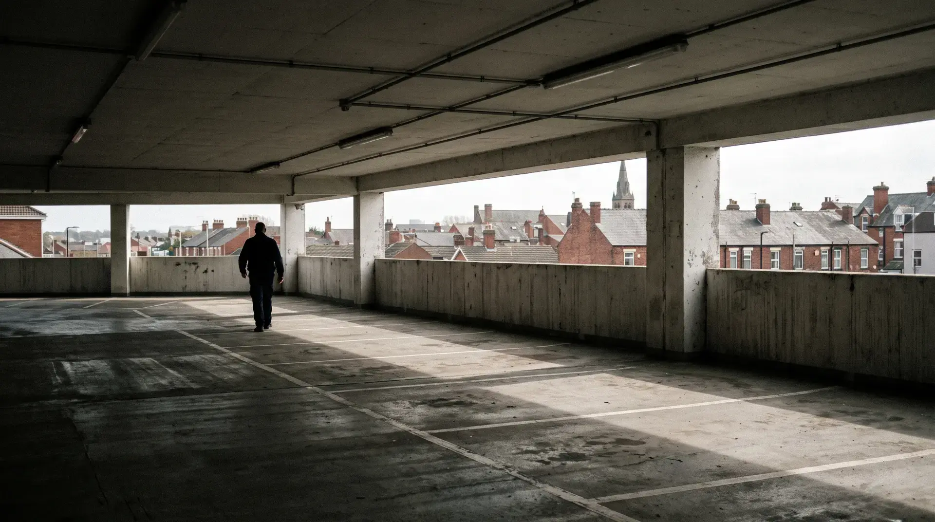 Lone security officer walking the empty upper deck of a British multi-storey car park on an overcast morning