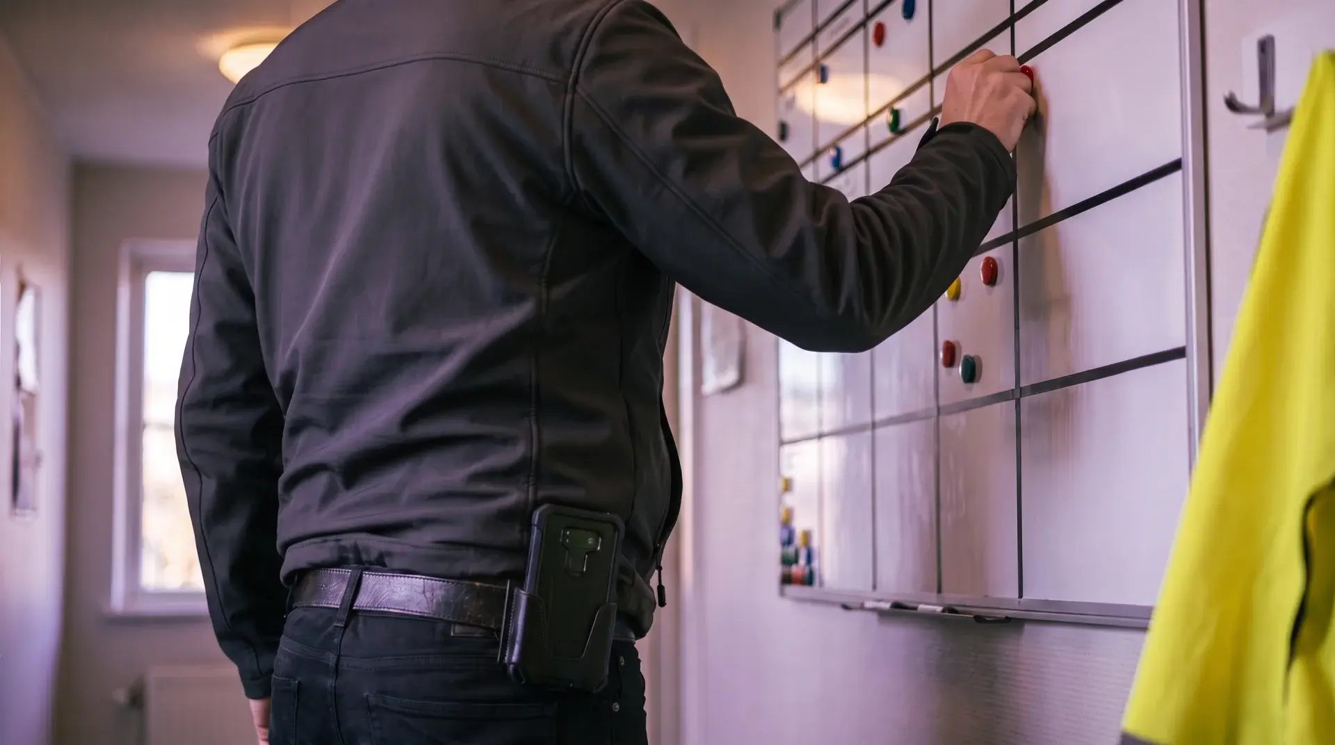 Einsatzleiter setzt im Betriebsbüro einen Magneten auf die Plantafel, Diensthandy im Gürtelholster, Warnweste am Haken