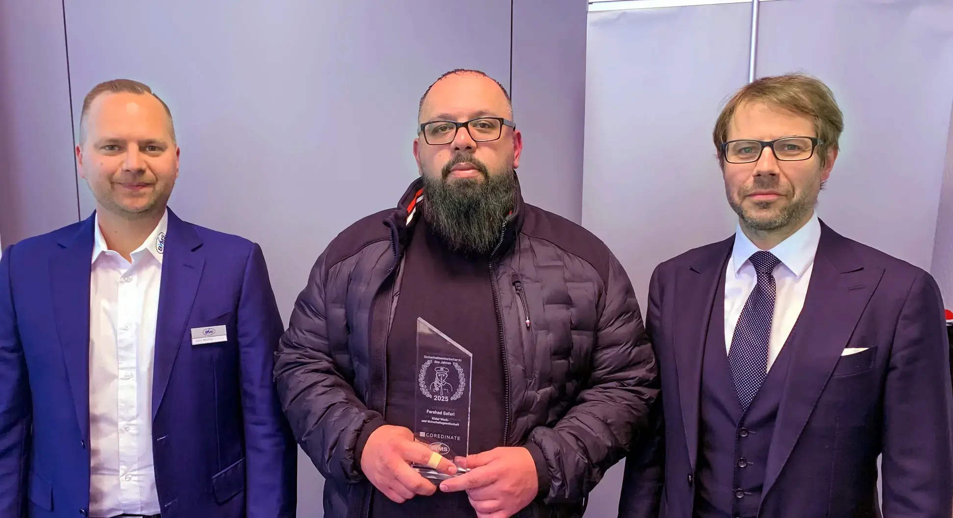 Farshad Safari stands between Lars Müller (left) and Michael Kulig (right) with his award for “Sicherheitsmitarbeiter des Jahres 2025” and holds the trophy in his hands.